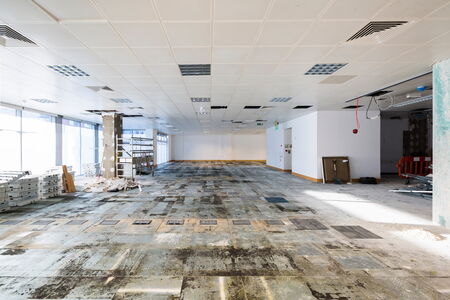 Großraumbüro während der Entkernung: beschädigter Bodenbelag, freiliegende Kabel, abgehängte Decke mit offenen Modulen und Baustellenmaterialien wie Gerüst und Kartons.