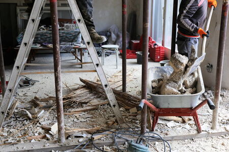 Baustellenszene im Innenbereich: Eine Schubkarre mit Bauschutt, ein Bauarbeiter mit Schaufel, herumliegender Putz und Holzlattenreste, eine Leiter sowie Stützen – deutliche Spuren der Entkernung und Rückbauarbeiten.