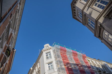 Blick von unten auf ein Altbaugebäude mit eingerüsteter Fassade in der Stadt, umgeben von weiteren klassischen Häusern, unter wolkenlosem blauen Himmel